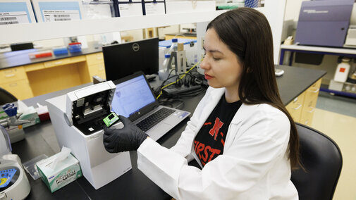 A woman with long dark hair and wearing a lab coat sits at a lab bench. She is wearing black gloves and holding a small cartridge in her left hand. Behind her, on the bench, is an open laptop, a plate reader. In the background of the image other lab benches with instruments including a flow cytometer can be seen.