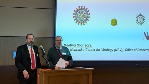 Two men standing at a podium. One is holding a paper. Both are looking at the camera and smiling. Behind them is a screen which says welcome and has images of viruses.