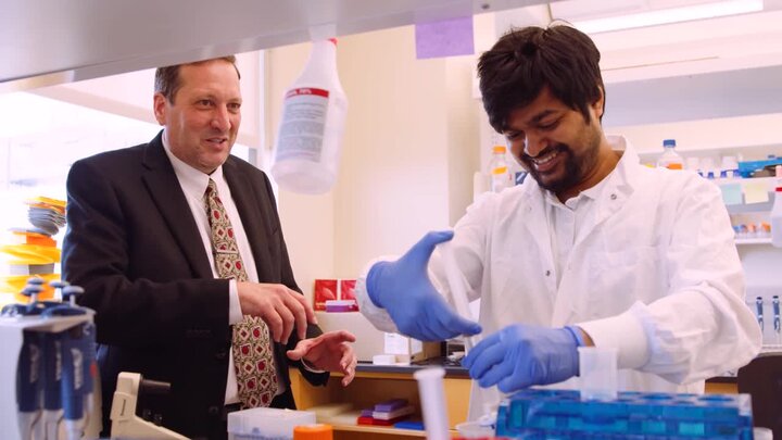 two people, one in a suit and one in a white lab coat and blue gloves, stand a lab bench. Both are happy.