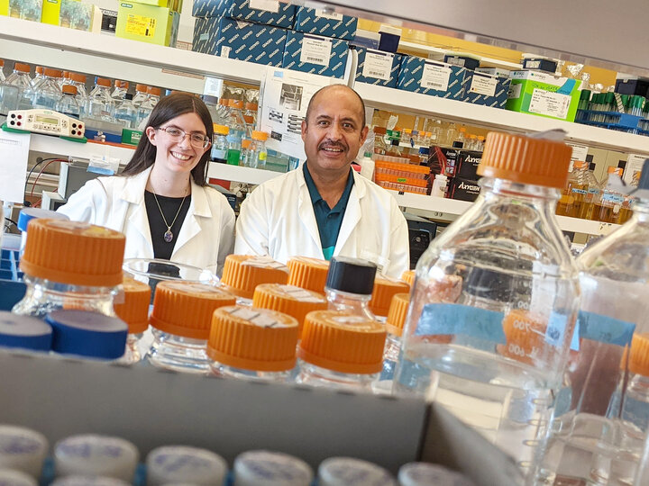 Two people stand in a lab. The image is from behind a set of shelves. In the foreground are glass bottles with orange tops. Behind the people are more lab benches with boxes and equipment. On the left is a woman with dark hair , glasses, and a necklace wearing a black shirt. On the right is a man with a mustache and teal shirt. Both people are wearing lab coats, smiling, and are visible above the waist behind the bottles. 