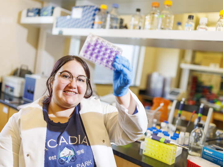 A woman with glasses and dark hair pulled back is standing in a lab. She is wearing a blue t-shirt and an open lab coat with blue gloves. She is looking up at a plastic well plate stained with purple dye that she is holding in her left hand. Behind her and slightly out of focus is a molecular lab bench with equipment, pipettes, and supplies.