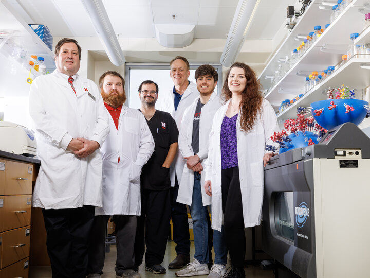 Six people stand in a semi-circle in a lab. To the left and right of the people are lab benches with equipment including centrifuges and shelves with lab plastics. On the right side of the image is a large model of a virus on display. Five people are wearing lab coats and one is in scrubs. All people are smiling and looking down at the camera.
