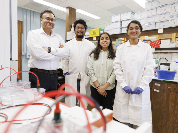 A group of four people stand in a lab. In the foreground are gel electrophoresis chambers. In the background are shelves with boxes of lab supplies. The people stand in a shallow semi-circle from left to right a dark haired man with glasses, a white shirt, and black pants with his arms crossed and watch visible. To his left is another man with glass and a beard wearing a white lab coat. Next to them are a women with dark hair over her shoulders wearing a light colored hoodie and another woman with her hair 