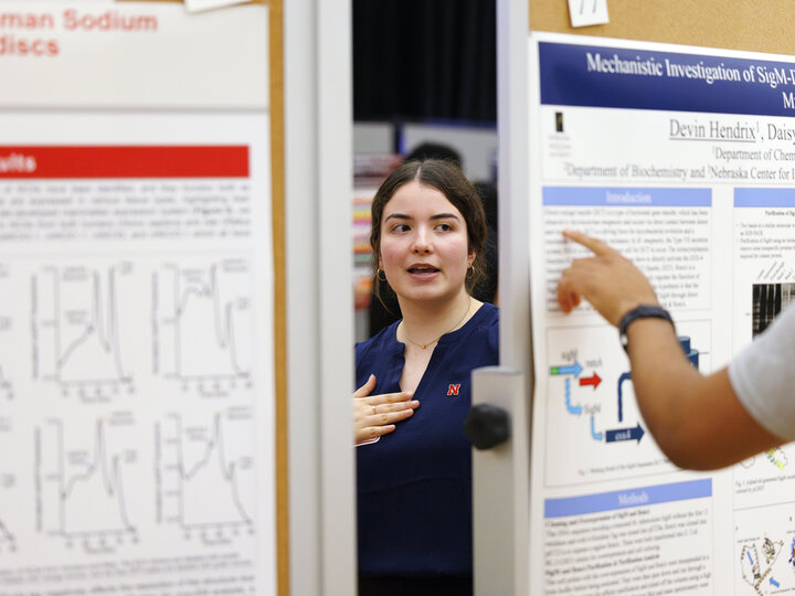 A young woman with dark hair pulled back in a blue shirt with a red letter N pin is standing between two scientific posters and discussing her work with an unseen audience.
